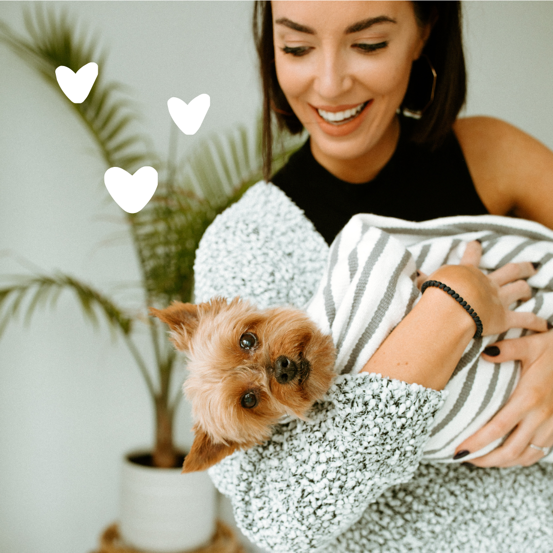 Woman holding a small dog wrapped in a striped blanket with heart graphics.